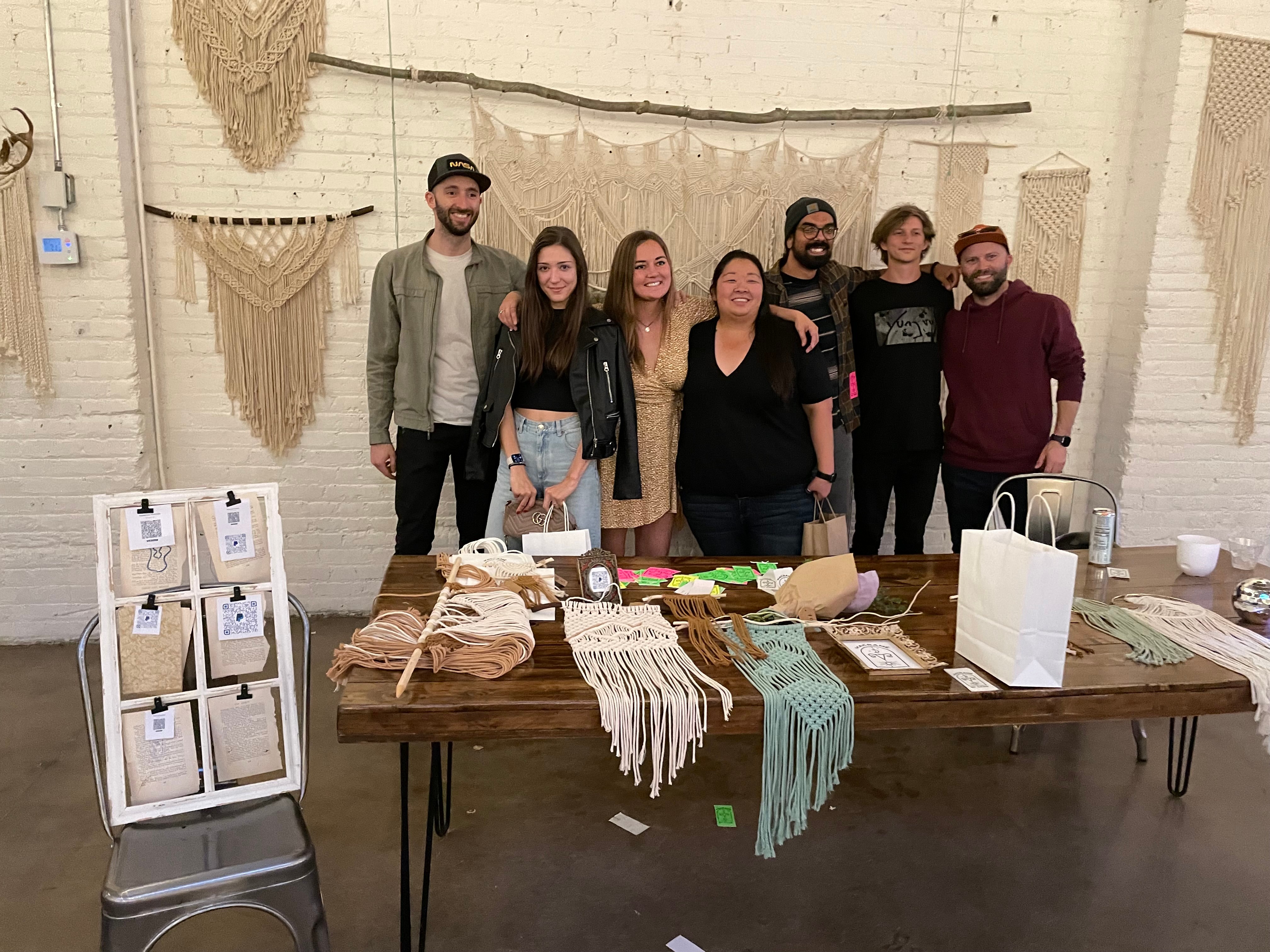 A group of makers gathered around a table of handmade macramé pieces at a market